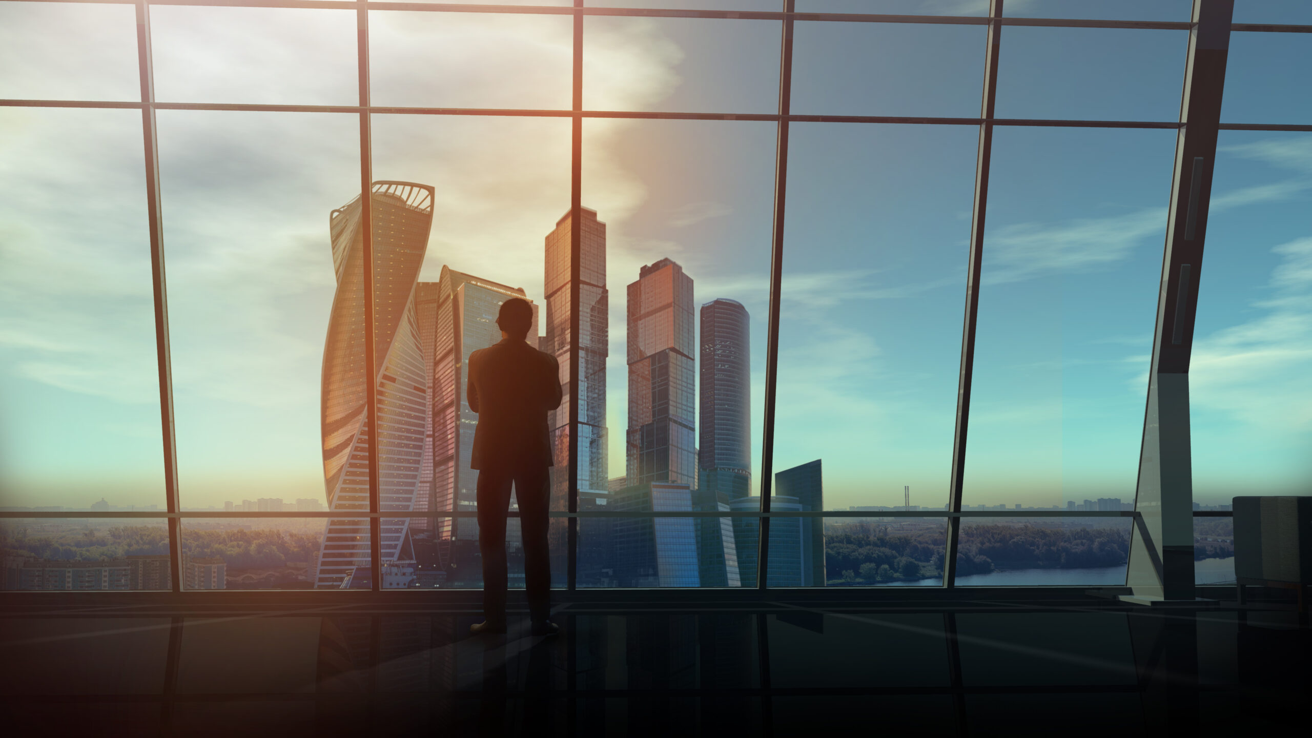 A businessman is standing in his office against the backdrop of a huge window overlooking skyscrapers.